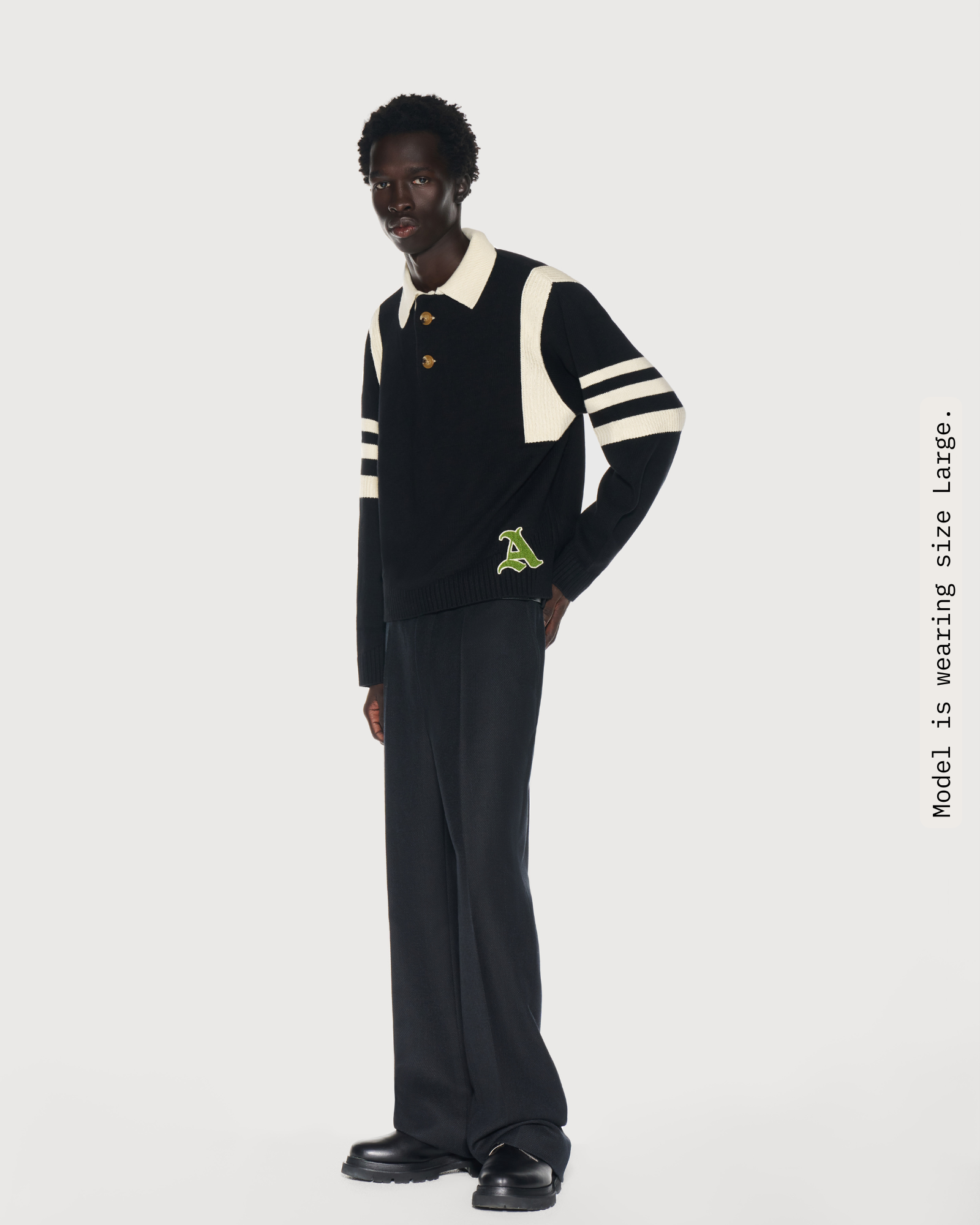 Man wearing a black rugby knit sweater with cream stripes and a green chenille logo. Aseye''s favorite sweater, the Kai Rugby Kni, was  inspired by the designer's brother, Kyrie Irving. 