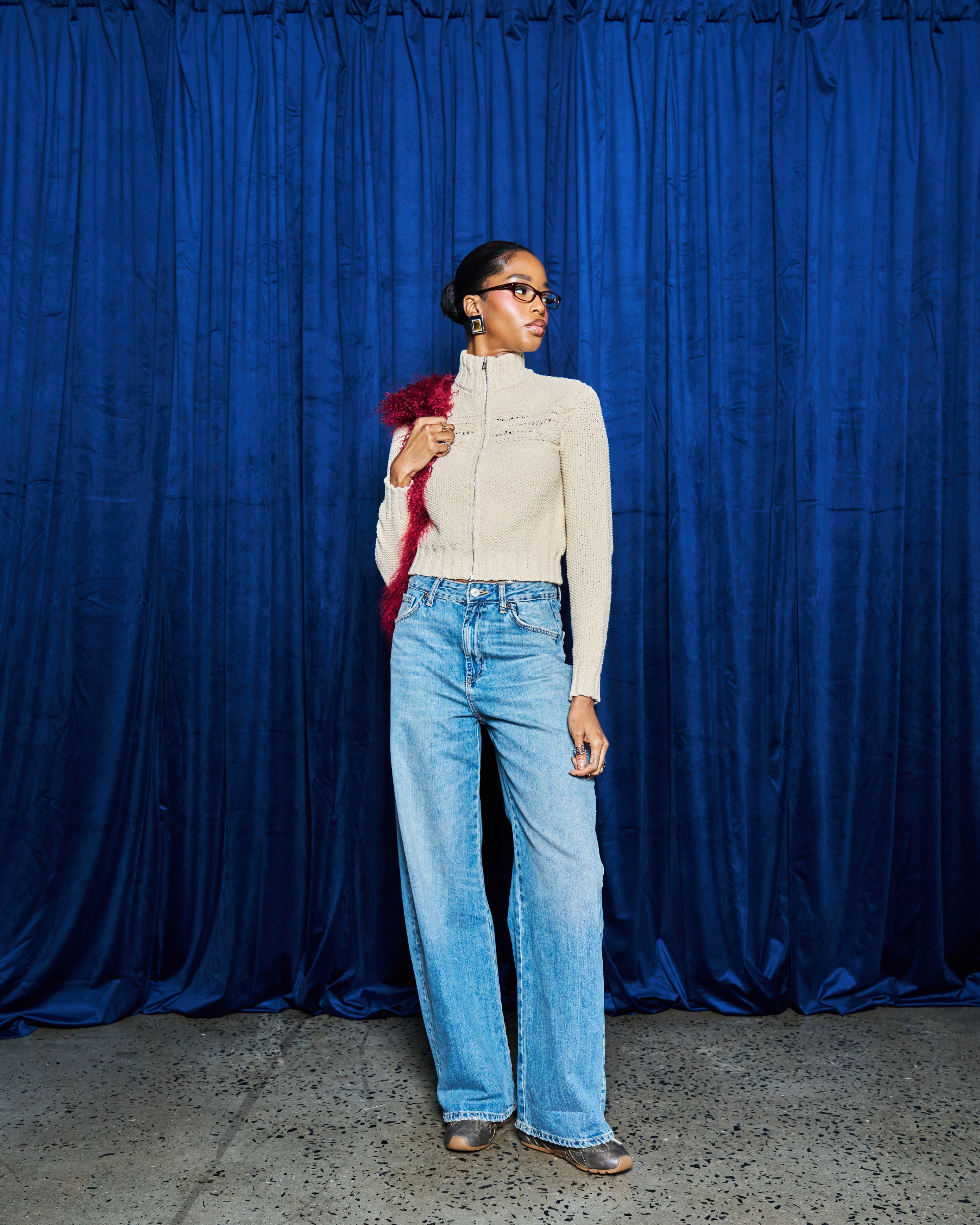 Person wearing a beige sweater and light blue jeans against a blue curtain background