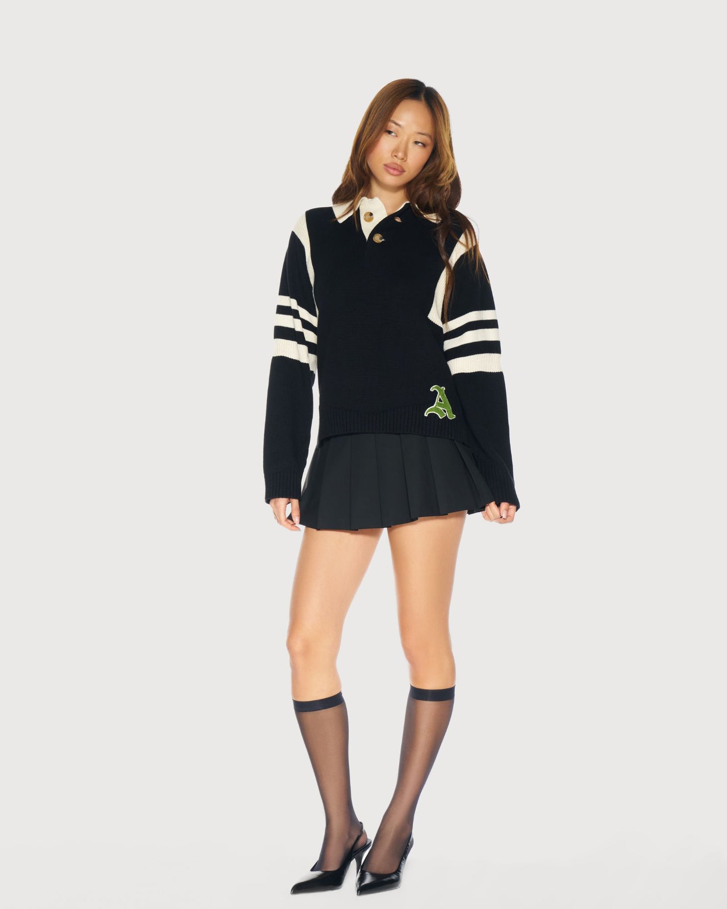 Woman wearing a black sweater with cream stripes and green chenille logo; paired with a skirt on a plain background